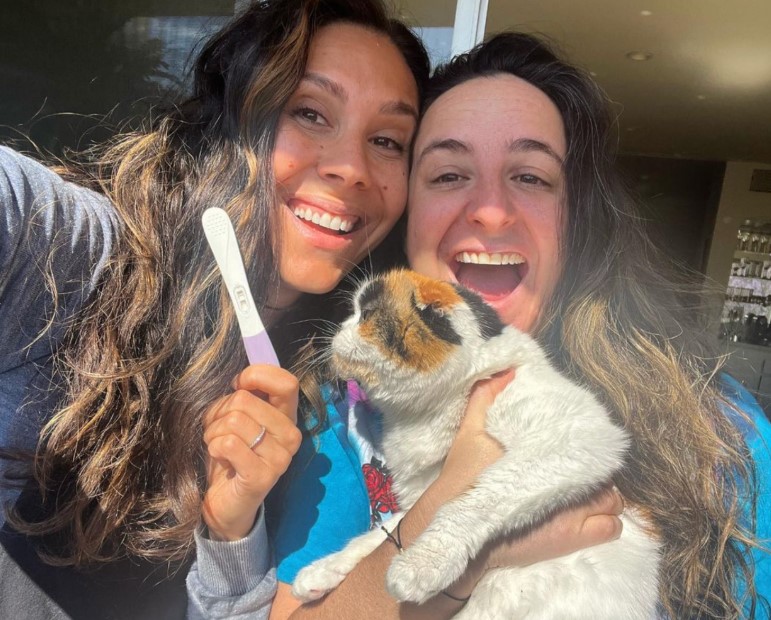 Clare Wasserman and her wife happily smiling showing a pregnancy test to the camera
