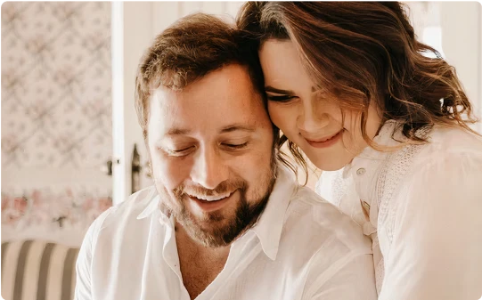 A couple smiling and looking down together. The woman has her arms wrapped around him, resting her head on his shoulder